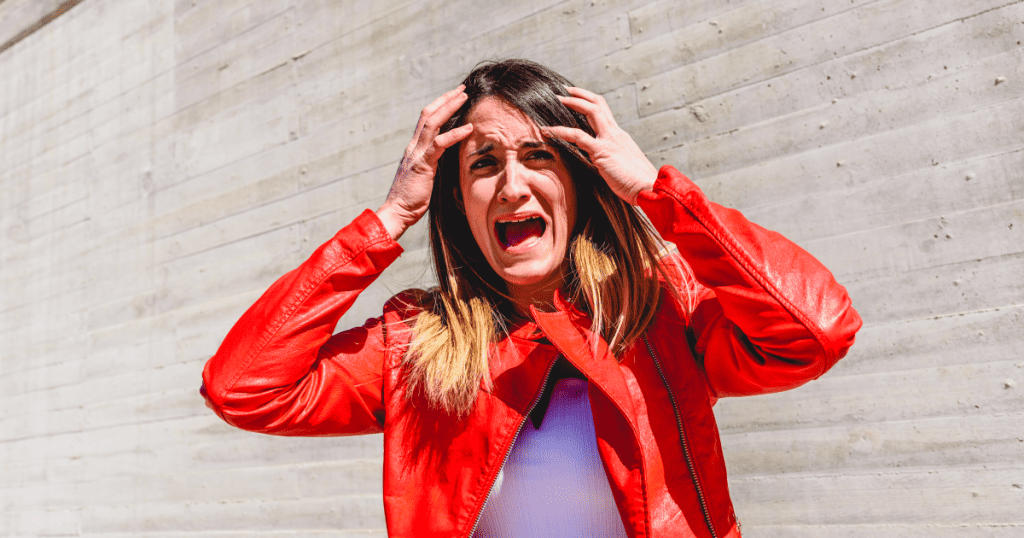 A woman in a red jacket clutches her head and screams in frustration while standing against a concrete wall, looking overwhelmed and furious.