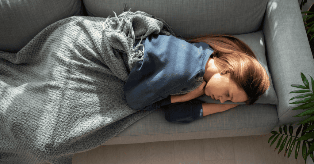 A drained woman lies curled on a couch under a blanket, capturing the deep exhaustion and loss of energy often felt after narcissistic abuse.