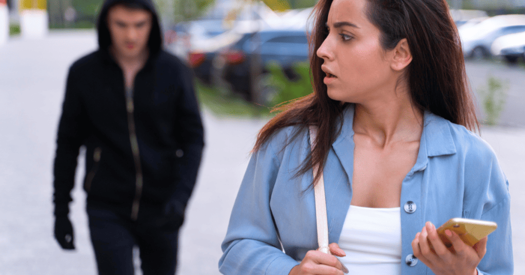 A woman walking outdoors looks over her shoulder with concern while holding her phone, symbolizing how narcissists secretly monitor or track their victims even after separation.