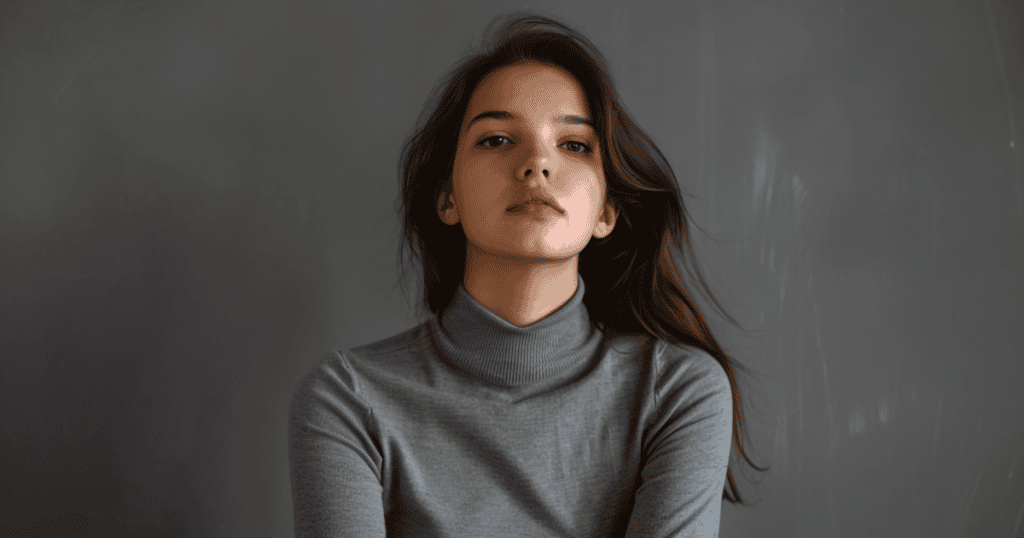 A young woman in a gray turtleneck sits against a neutral gray background, facing the camera with a calm, composed expression and steady posture.