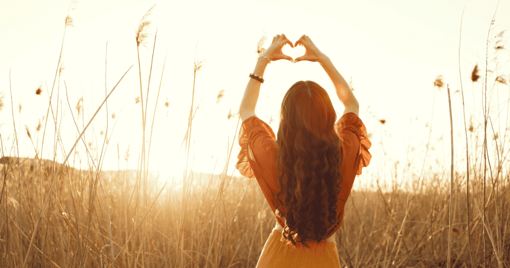 A woman stands in a sunlit field with her back to the camera, lifting her arms to form a heart shape with her hands as warm golden light shines around her.