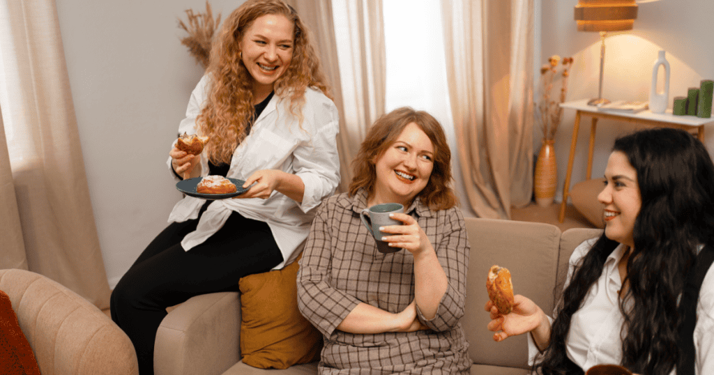 Three women laugh together on a couch while one leans in with a pastry, showing someone inserting themselves into a joyful moment to steal attention.