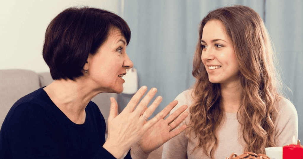 An older woman gestures dramatically while speaking to a polite younger relative, dominating the conversation to ensure all eyes remain on her.