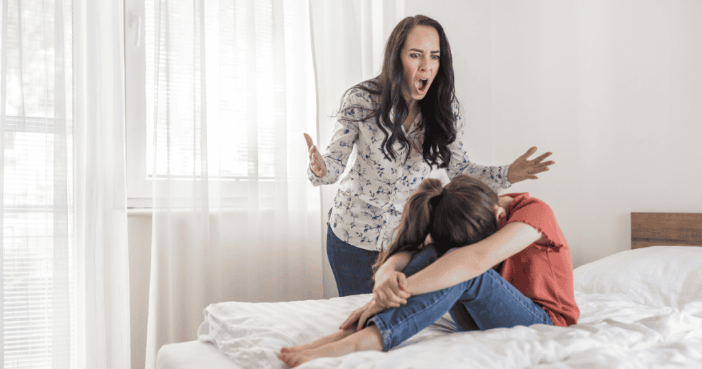 A woman sits curled up on a bed while another woman stands over her yelling, illustrating how narcissists often pick fights before bedtime to keep others emotionally off-balance and exhausted.