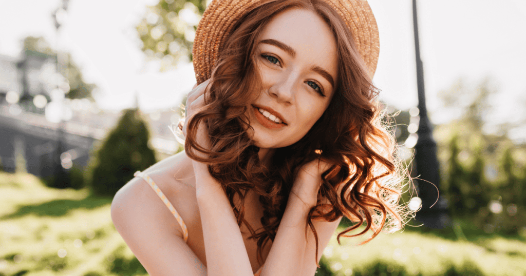 A smiling, attractive young woman with long curly hair and a straw hat poses outdoors, indicating a visual state that could trigger a negative response.