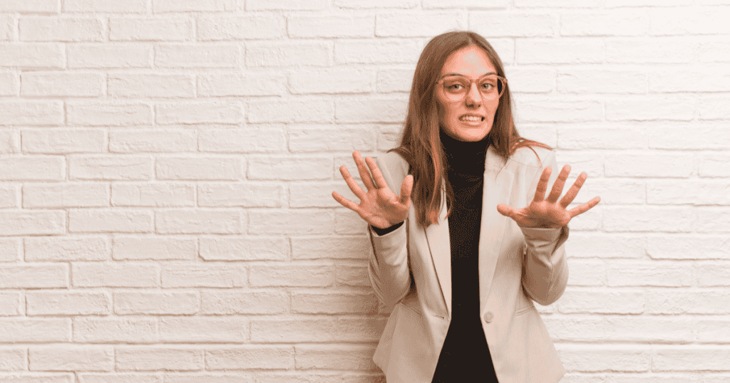 A woman stands against a white brick wall with her hands raised in a startled, uneasy gesture, capturing the confused realization people have when noticing strange narcissist behaviors that suddenly don’t feel normal.