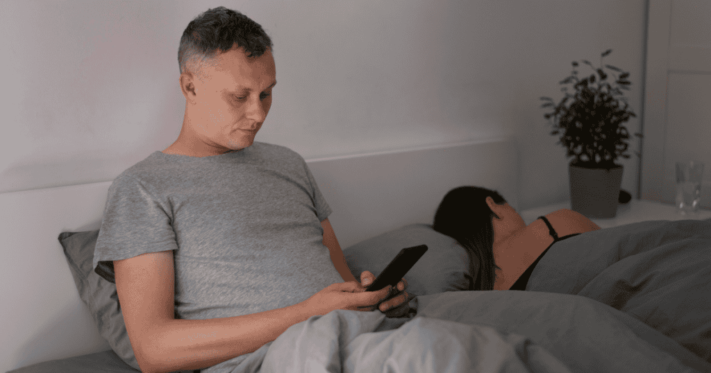 A man lying in bed looks at his phone while his partner sleeps beside him, highlighting a new need for digital privacy and late-night secrecy.
