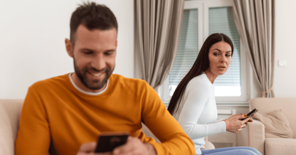 A woman sits holding her phone and looking uneasy while her partner smiles at his phone beside her, suggesting silent infidelity and hidden behavior in a narcissistic relationship.