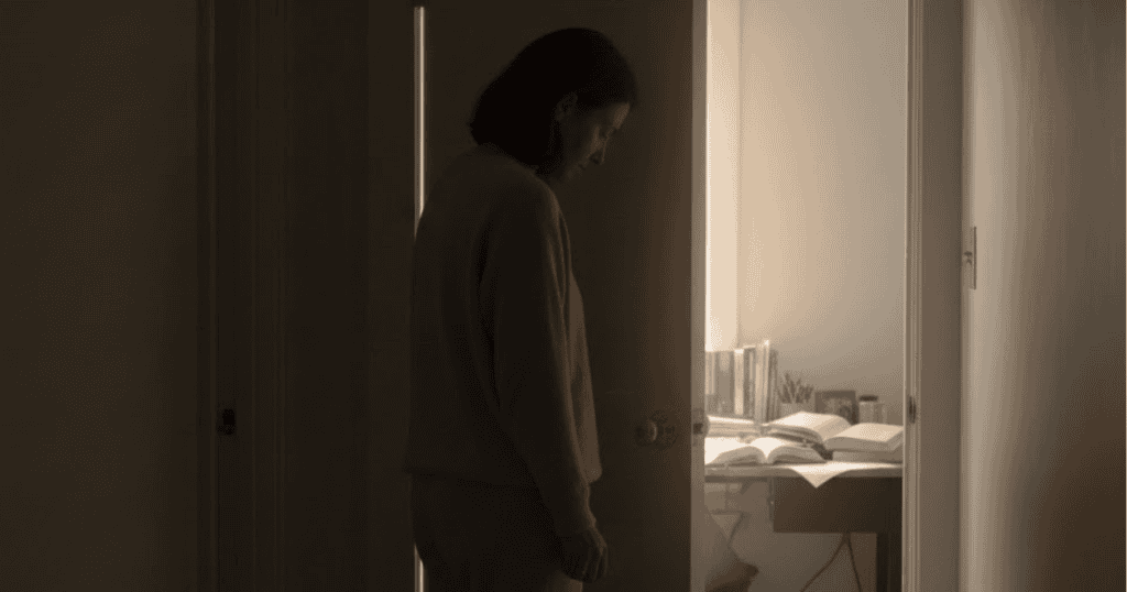 A woman is silhouetted in a dark doorway looking into a sunlit room with open books on a desk, hinting at a private life that is carefully kept separate from public view.