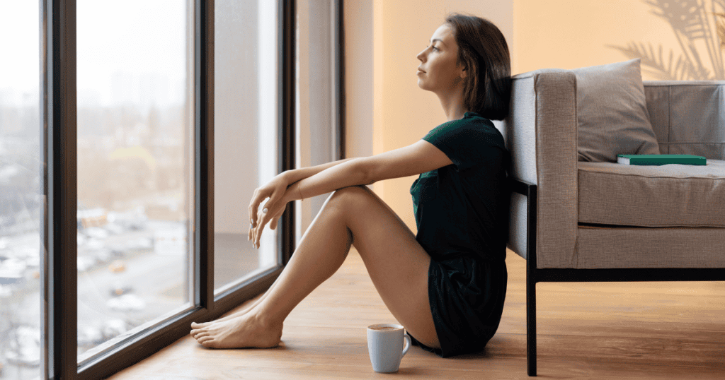 A woman sits quietly on the floor by a large window with a cup of coffee, reflecting deeply on the true nature of her surroundings.