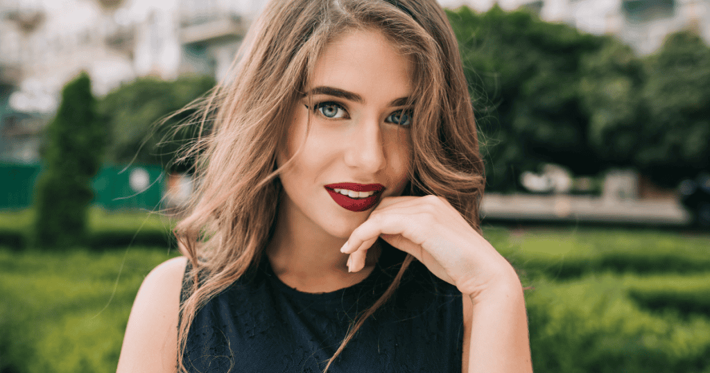 A beautiful young woman with long wavy hair and red lipstick smiles over her shoulder outdoors, highlighting a natural brilliance that cannot be taken.