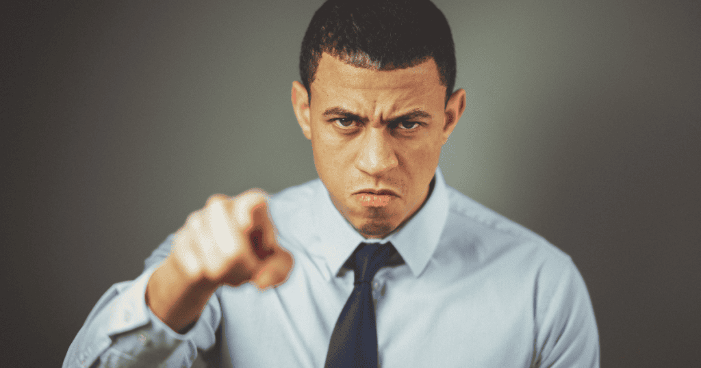 A man in a professional shirt and tie glares intensely while pointing his finger directly at the camera, capturing the confrontational essence of a personality that exerts control through intimidation.