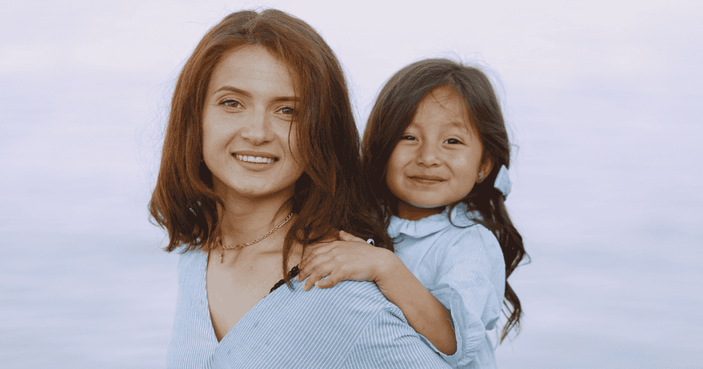 A smiling woman gives a young girl a piggyback ride against a soft background, representing a tender reconnection with a past identity.