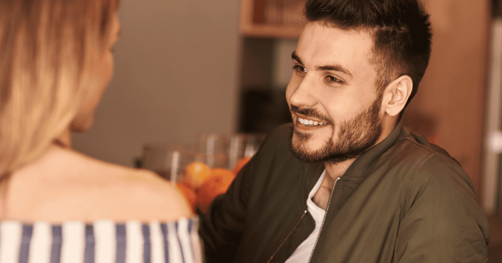 A smiling man leans in to listen to his date, his charming expression hiding a calculating attempt to assess her character.