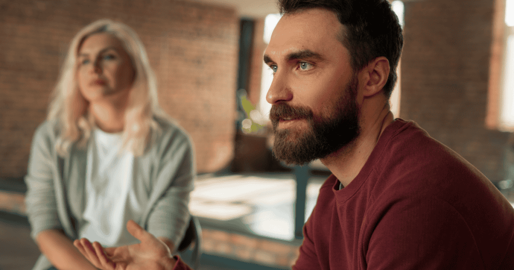A bearded man gestures earnestly in the foreground while a woman sits calmly in the blurred background, illustrating the quiet strength of emotional detachment.