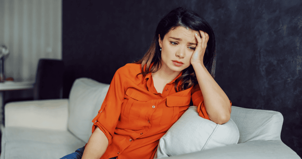 A distressed woman sits on a sofa holding her head in her hand, physically mirroring the exhaustion of constantly questioning her own judgment.