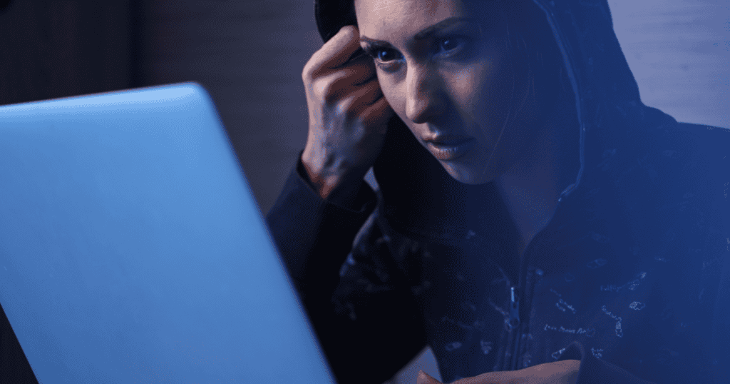 A woman in a dark hoodie stares intently at a glowing laptop screen in a dim room, her focused expression hinting at a private, nocturnal fixation.