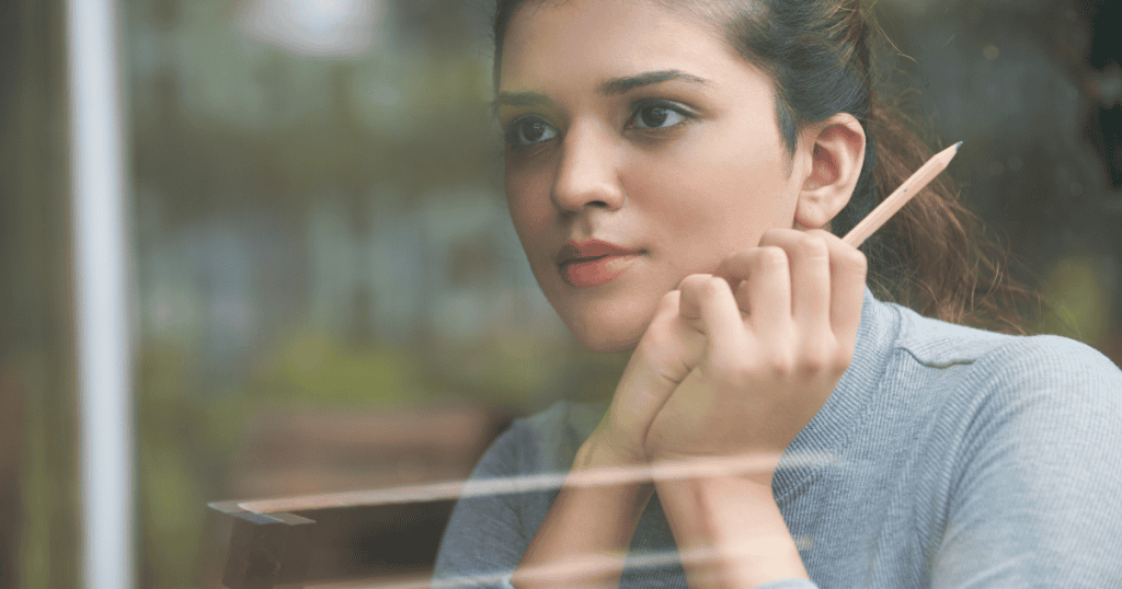 A contemplative woman holds a pencil while looking out a window, reflecting the internal resolve needed to define personal limits.