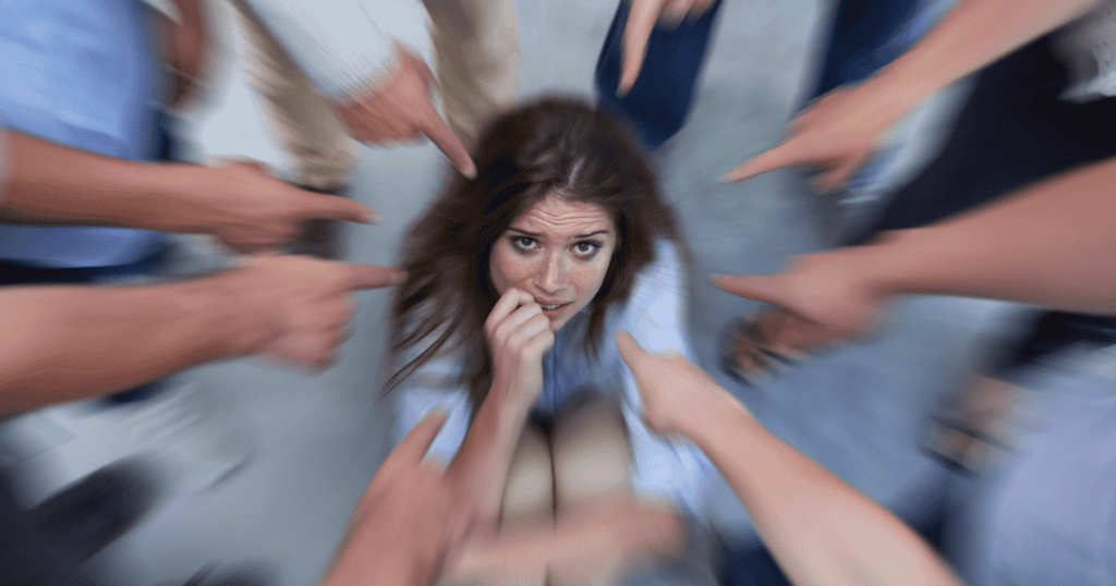 A fearful woman crouches on the floor as blurred fingers point accusations from every angle, illustrating the rapid escalation of blame that occurs in volatile environments.