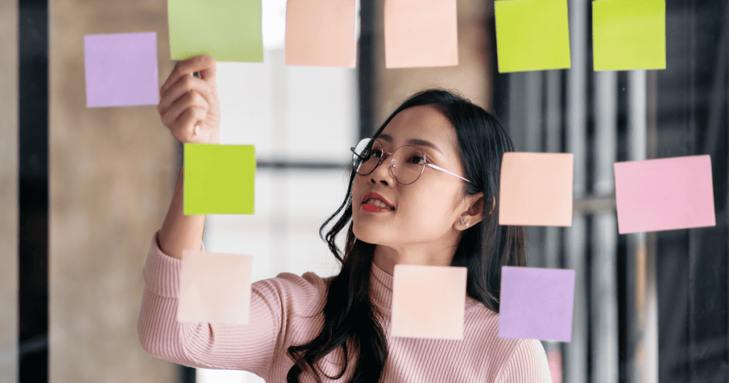 A focused woman organizes sticky notes on a transparent board, representing the clarity and strength that comes from building a strategy rather than reacting to chaos.