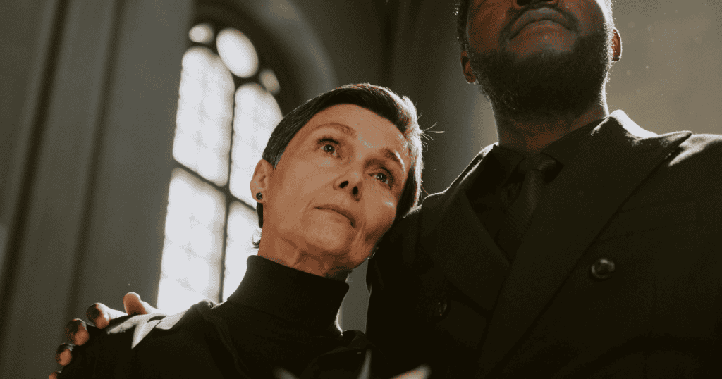 An older woman leans sorrowfully against a companion in a church, her upward gaze appearing more like a performed plea for attention than genuine mourning.