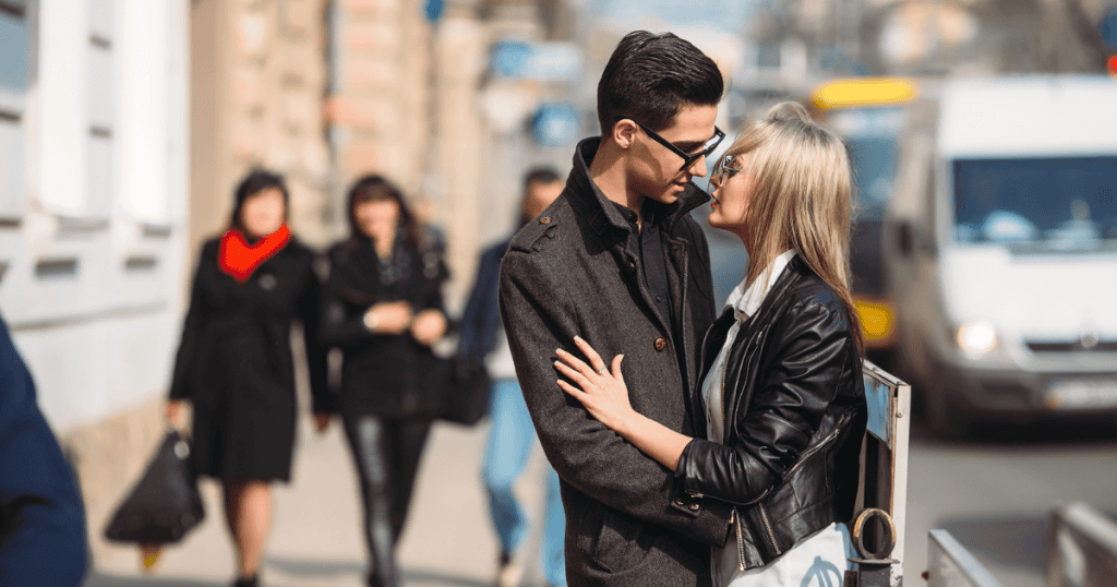 A couple standing close on a busy city sidewalk, performing an embrace that seems more intended for the surrounding audience than for themselves.