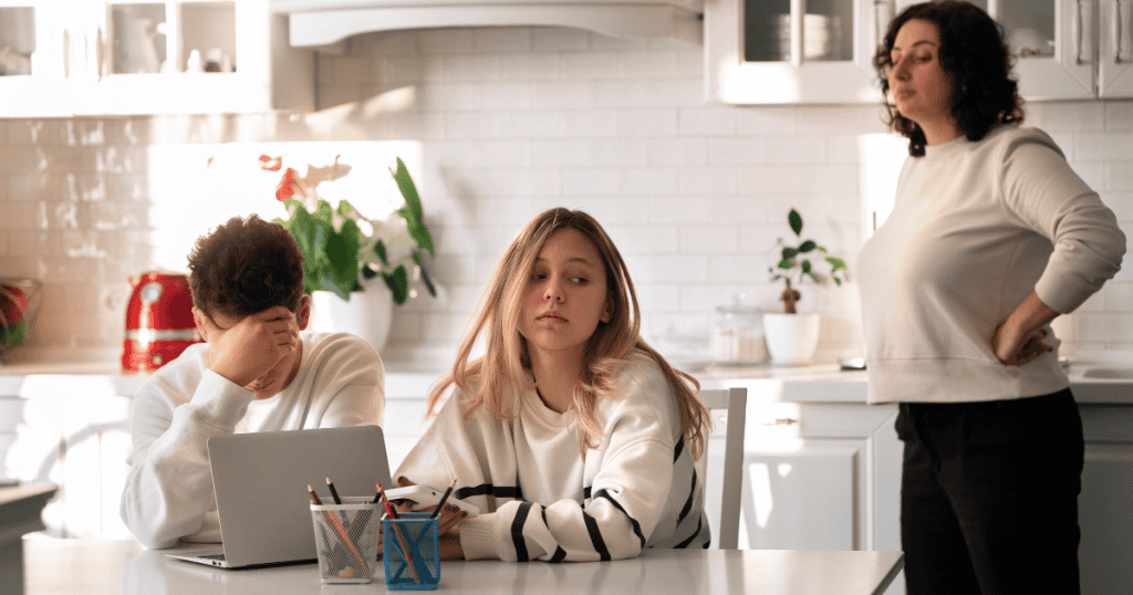 Two teenagers sit tensely at a table while a woman stands behind them with hands on her hips, capturing a household defined by underlying instability.