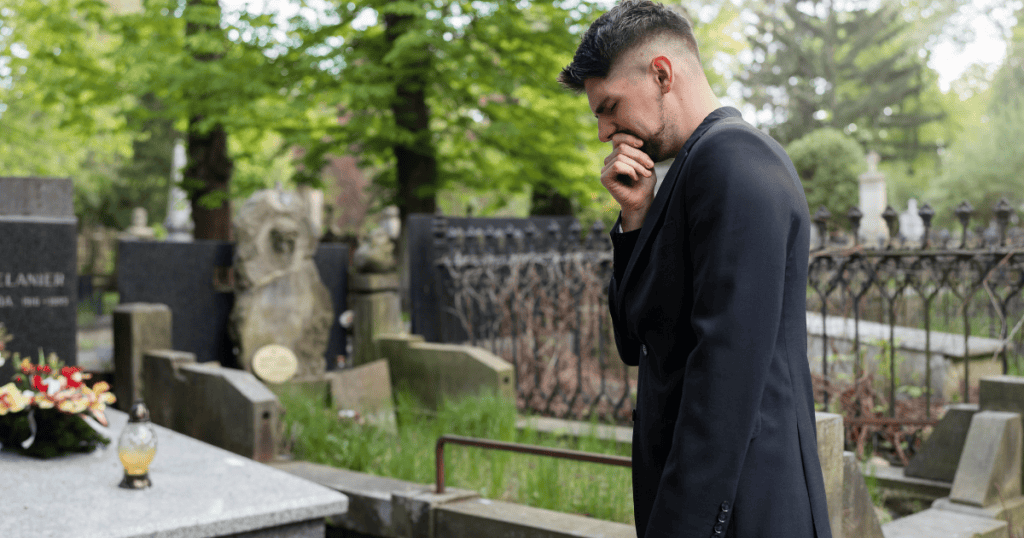 A man stands beside a tombstone with his hand covering his mouth, his pose looking too rehearsed to be an authentic moment of reflection.
