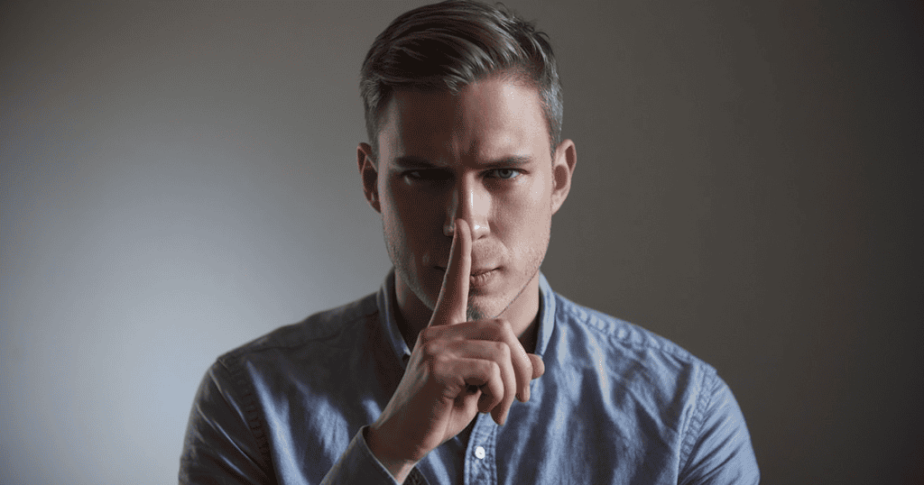 A man holds a single finger to his lips against a dimly lit background, embodying a calculated refusal to disclose what remains hidden.