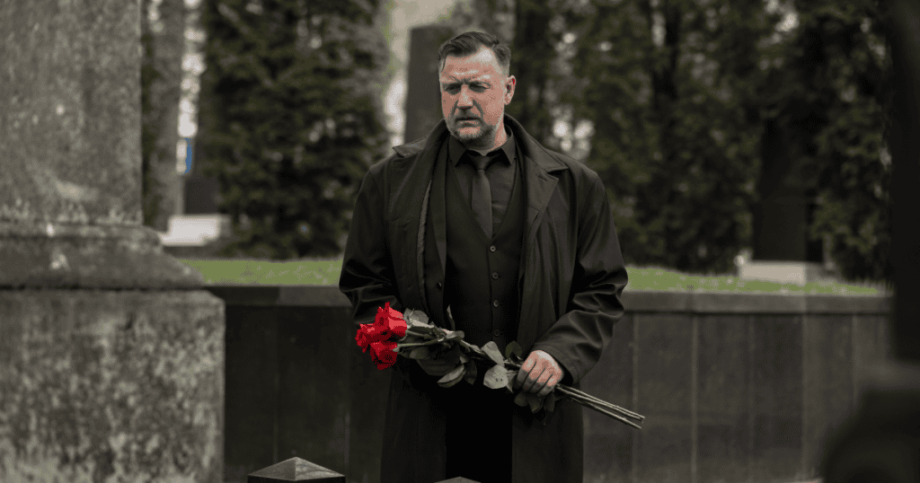 A stern man holds a bouquet of red roses while standing before a monument, his rigid posture commanding the spotlight rather than honoring the deceased.