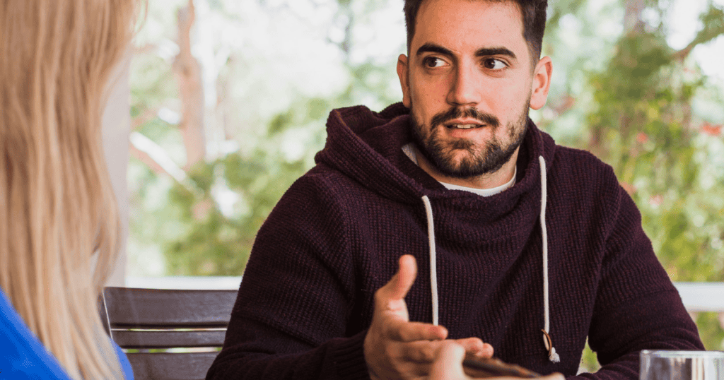 A man in a hoodie looks sideways while speaking to a companion, seemingly on the verge of admitting the uncomfortable and hidden envy driving his animosity.