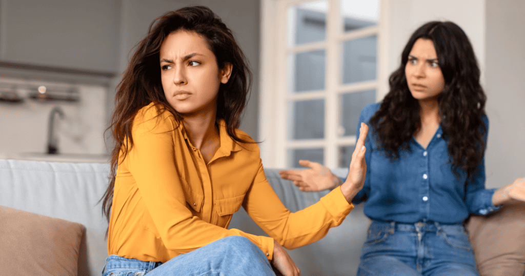 A woman sits on a couch with her hand raised in dismissal while another woman gestures toward her, illustrating avoidance of accountability during a tense confrontation.