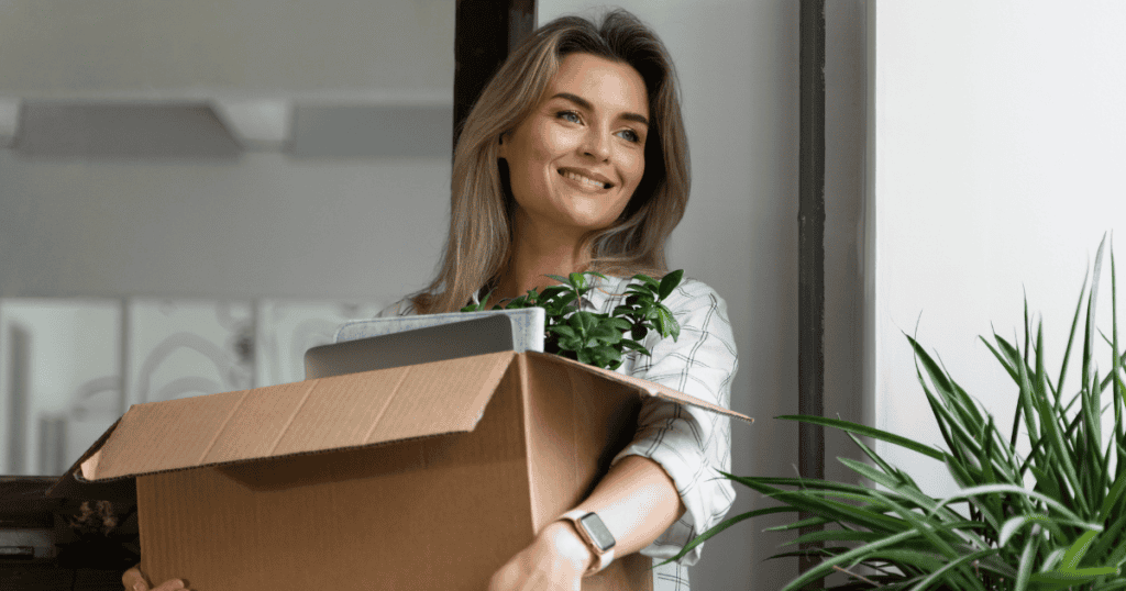 A smiling woman holds a packing box filled with belongings, symbolizing the difficult but liberating decision to physically separate from the past and move on.