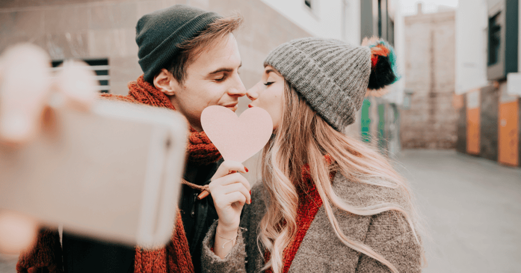 A couple dressed in winter clothes kisses behind a pink paper heart, capturing the warmth of a bond that feels safe rather than painful.