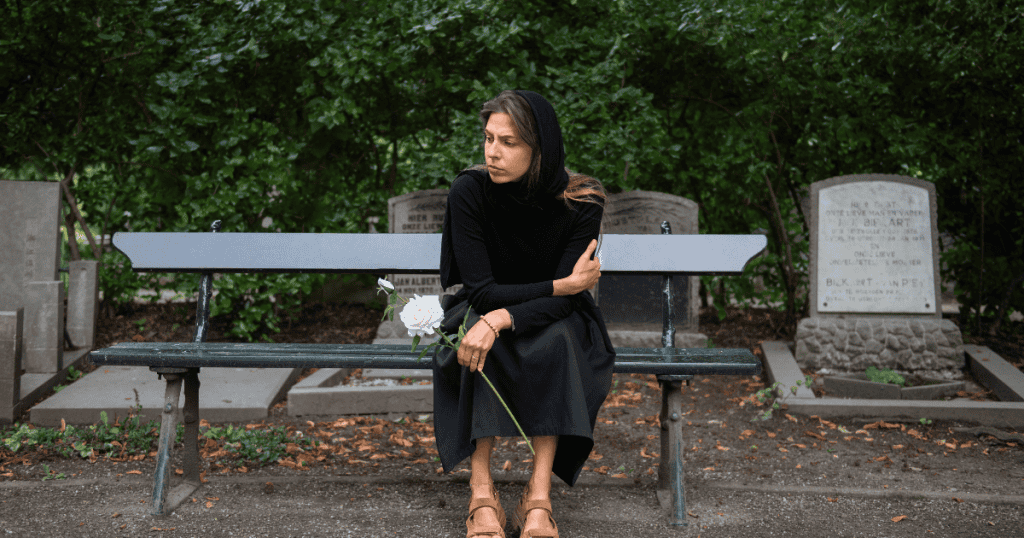 A woman sits isolated on a cemetery bench holding a single white rose, her guarded expression hinting at an inner attitude that the setting fails to mask.