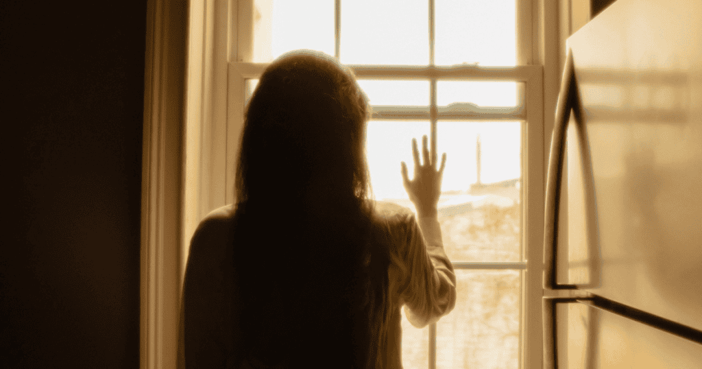A silhouette of a woman presses her hand against a window pane, staring out toward a light that she is only just starting to recognize.