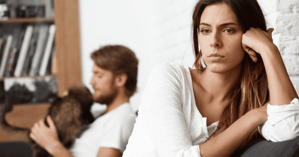 A woman looking away with a troubled expression while a man sits in the background, capturing the quiet isolation that can exist within a partnership.