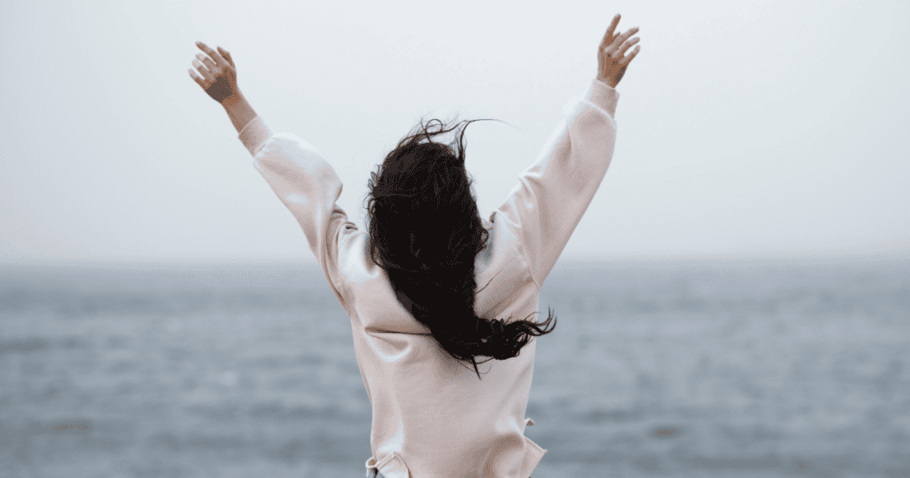 A woman stands facing the ocean with her arms raised wide, representing the liberation of a long-neglected inner spirit.
