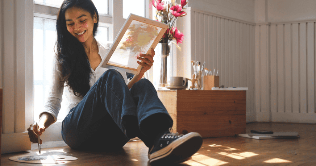 A woman sits on the floor bathed in sunlight while holding a canvas, focusing on the slow process of creating something new for herself.