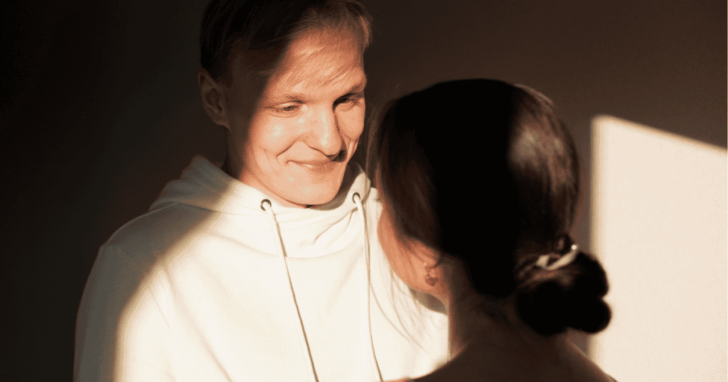 A man in a light hoodie smiles warmly at a woman standing in the shadows, capturing the moment a specific light draws someone in.