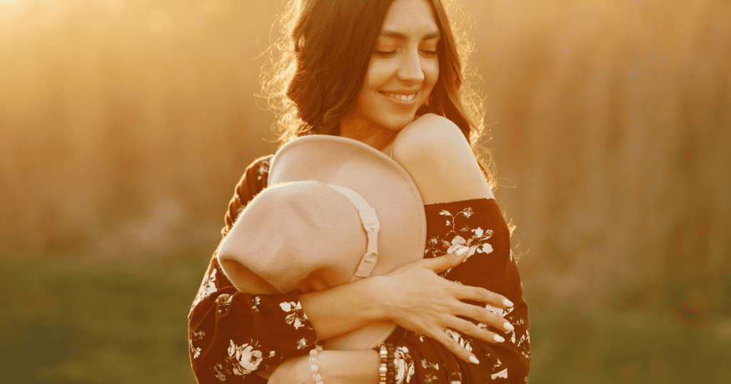 A smiling woman hugs herself in the golden light of sunset, finding peace in the release of carrying a negative burden that was never hers to hold.