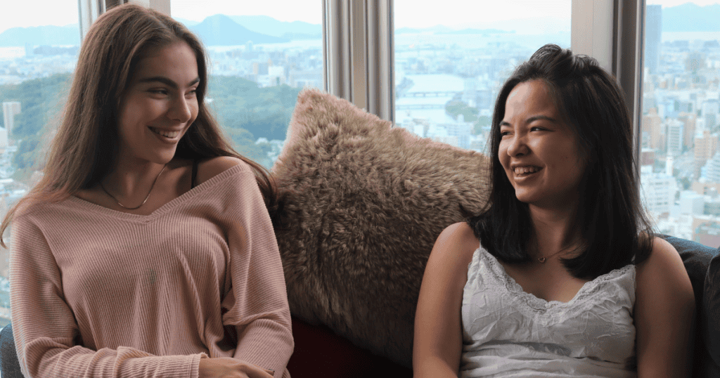 Two women sit close together on a sofa while laughing and talking, capturing a moment of intense focus and social validation.