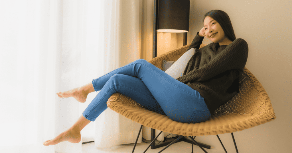 A woman reclines comfortably in a wicker chair near a bright window, her serene smile reflecting a newfound sense of personal peace.