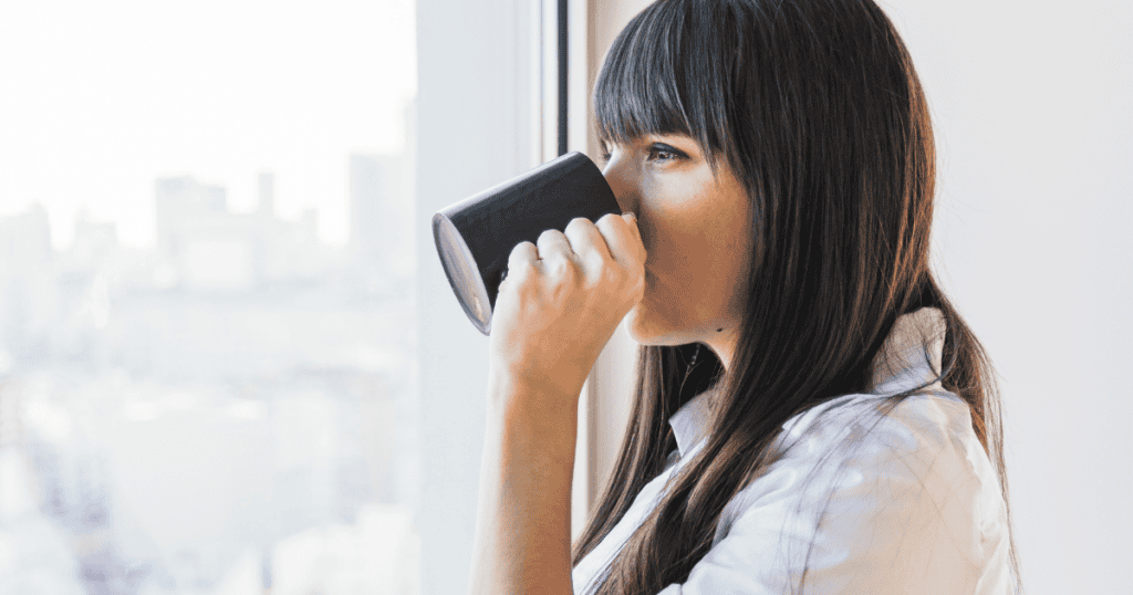 A woman looks out a bright window while sipping from a black mug, her quiet composure speaking volumes in the absence of conversation.