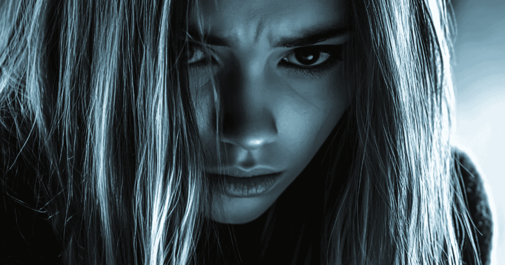 A tight, high-contrast close-up shows a woman’s face partially obscured by hair, her sharp and piercing stare reflecting a turbulent internal state.