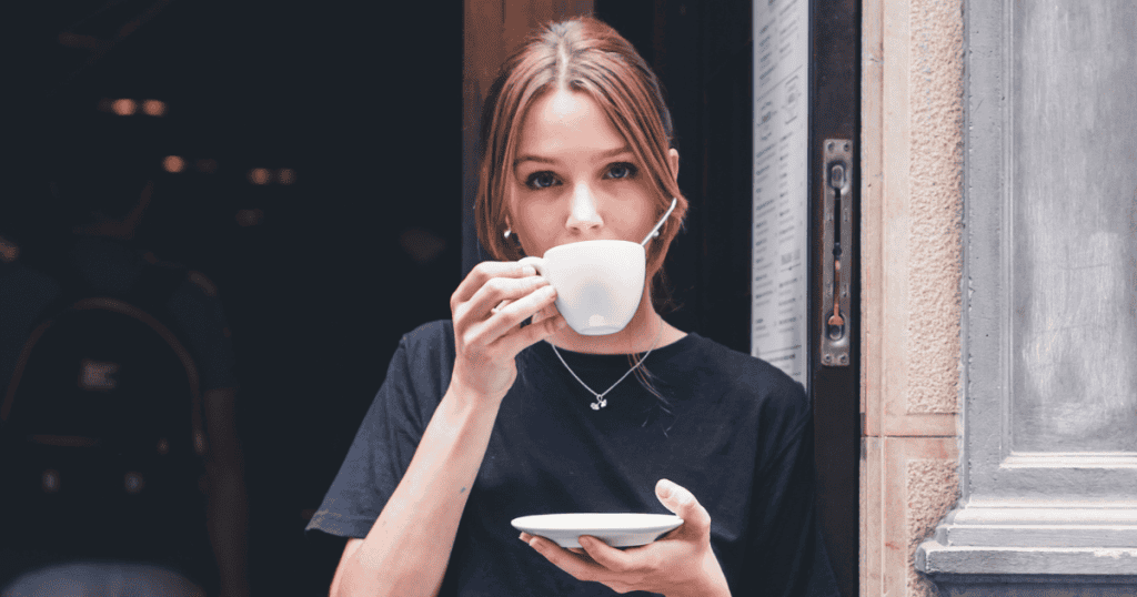 A woman peers over her coffee cup while standing in a doorway, her eyes widening as if silently reacting to an unsettling shift in the conversation.