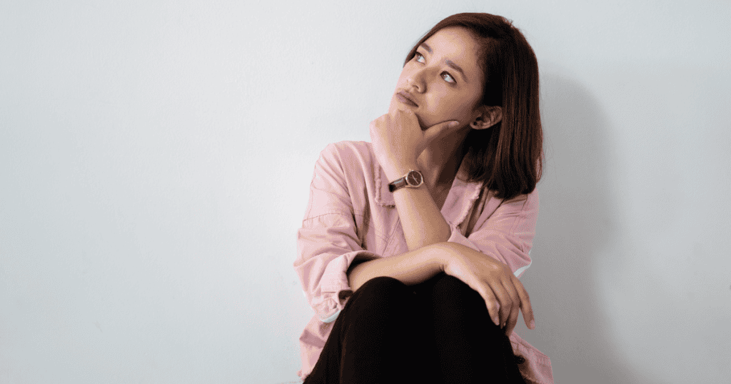 A woman sits against a plain white wall with her hand on her chin in deep thought, portraying the mental loop of questioning one's current position.