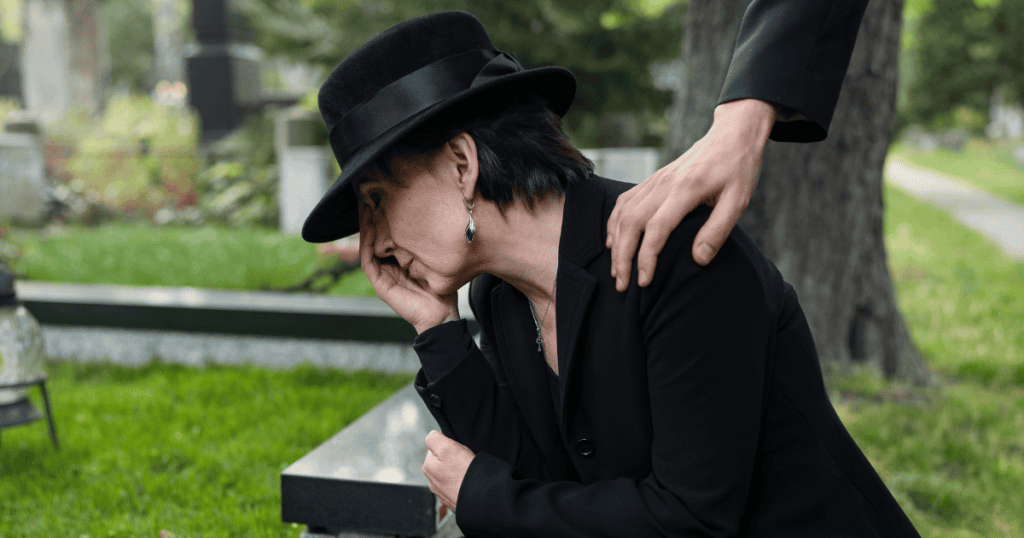A woman in mourning sits by a grave burying her face in her hand, her theatrical display of sorrow seemingly designed to elicit the comforting hand on her shoulder.