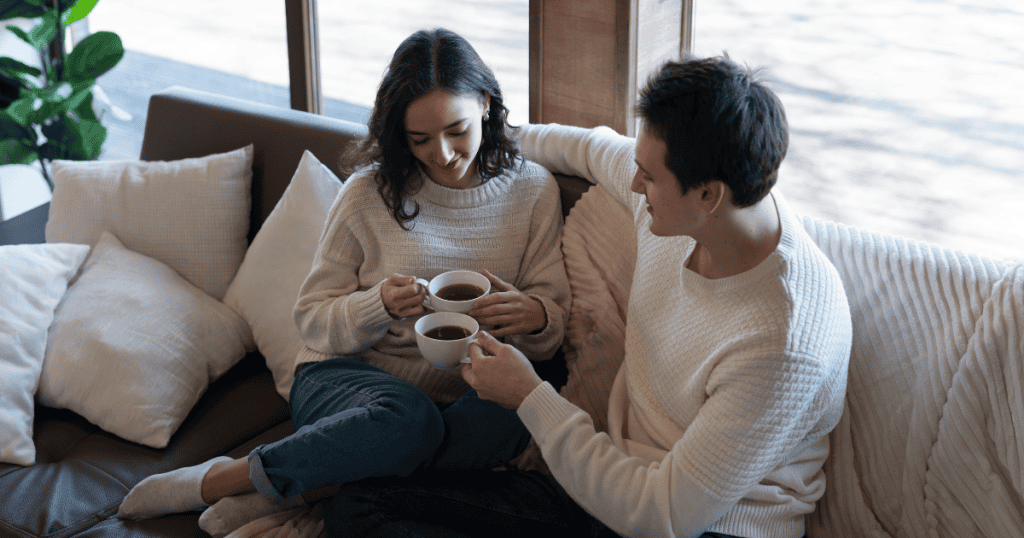 A man and woman relax on a sofa with warm mugs of tea, settling into a calmness that might initially feel surprisingly foreign to the nervous system.