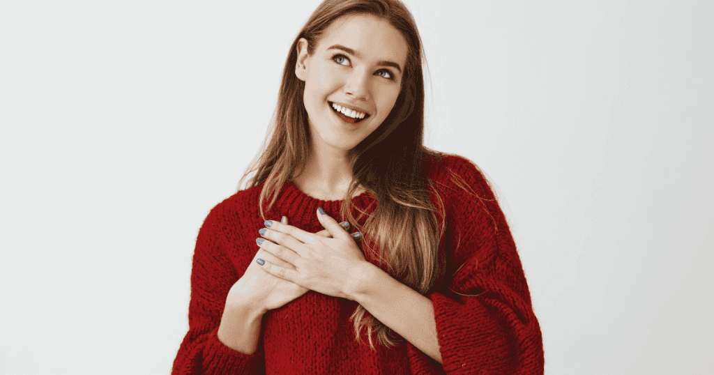 A joyful woman in a red sweater places her hands over her heart, visibly unburdened by the understanding that the mistreatment she faced was a result of circumstances beyond her control.